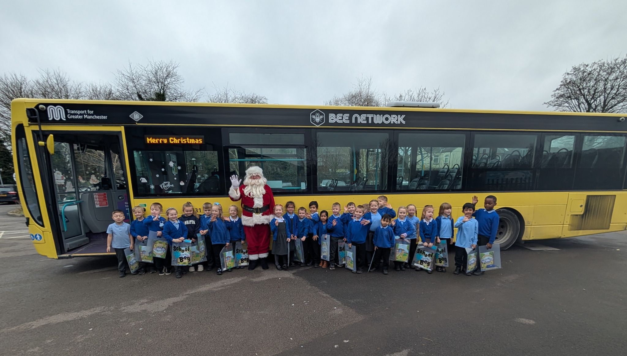 Santa standing with a group of young students waving in front of a yellow bus.